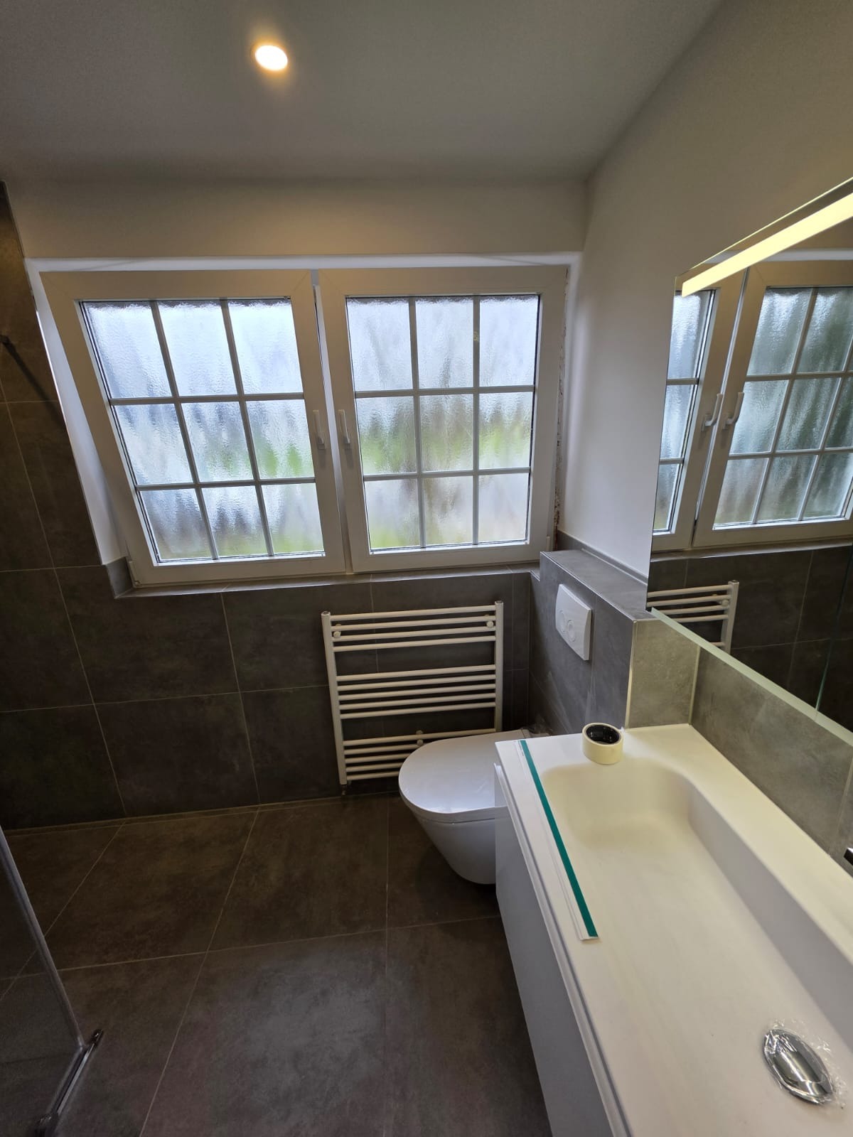 Nowoczesna łazienka z oknem dachowym i grzejnikiem drabinkowym. Szare płytki na ścianach i podłodze. Biała umywalka z lustrem z podświetleniem LED.