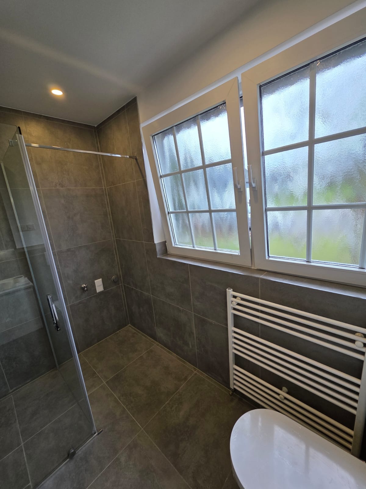 Nowoczesna łazienka z kabiną prysznicową typu walk-in, grzejnikiem drabinkowym i oknem z widokiem na zewnątrz. Szare płytki na ścianach i podłodze.