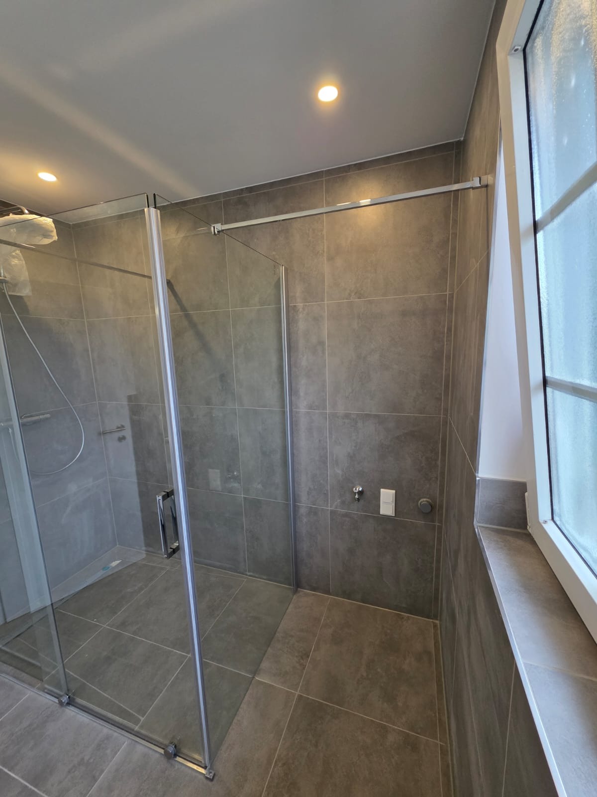 Nowoczesna kabina prysznicowa z szarymi płytkami w łazience. Widoczne szklane drzwi, minimalistyczna armatura i okno z białymi ramami. Czyste, eleganckie wykończenie.