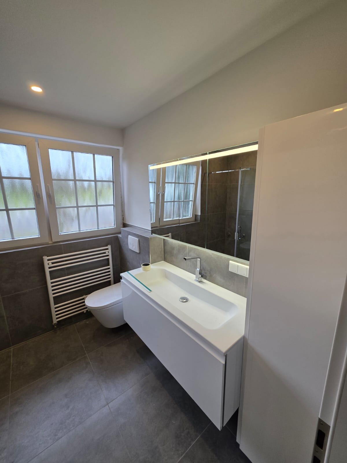 Nowoczesna łazienka z szarymi płytkami, białą szafką pod umywalką i dużym lustrem z podświetleniem LED. Widoczne okno z grzejnikiem.