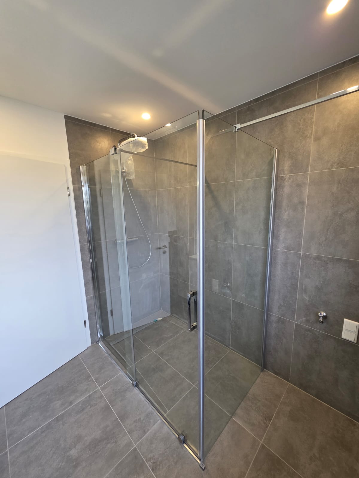 Szklana kabina prysznicowa z deszczownicą w nowoczesnej łazience wyłożonej szarymi płytkami. Widoczna biała ściana z drzwiami. Minimalistyczny design.