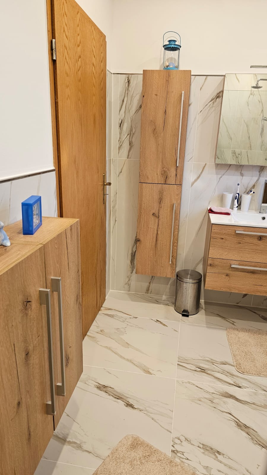 Jasna łazienka z marmurowymi płytkami, drewnianymi szafkami i wiszącą szafką z dekoracyjną latarenką. Umywalka z lustrem i metalowy kosz na śmieci.