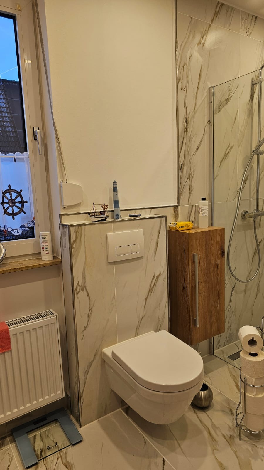 Nowoczesna łazienka z podwieszaną toaletą, marmurowymi płytkami i akcentami marynistycznymi na parapecie. Umywalka z szafką z drewna, kabina prysznicowa za toaletą.