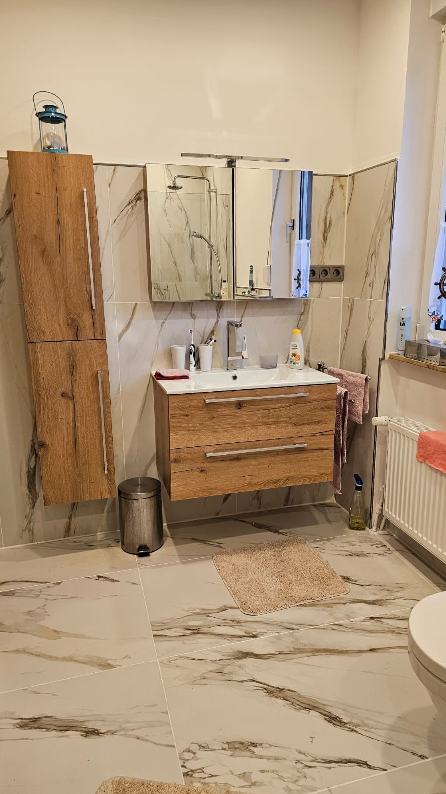 Jasna łazienka z drewnianą szafką pod umywalką, marmurowymi płytkami i wiszącą szafką z lampionem na górze. Umywalka z baterią i kosmetykami.