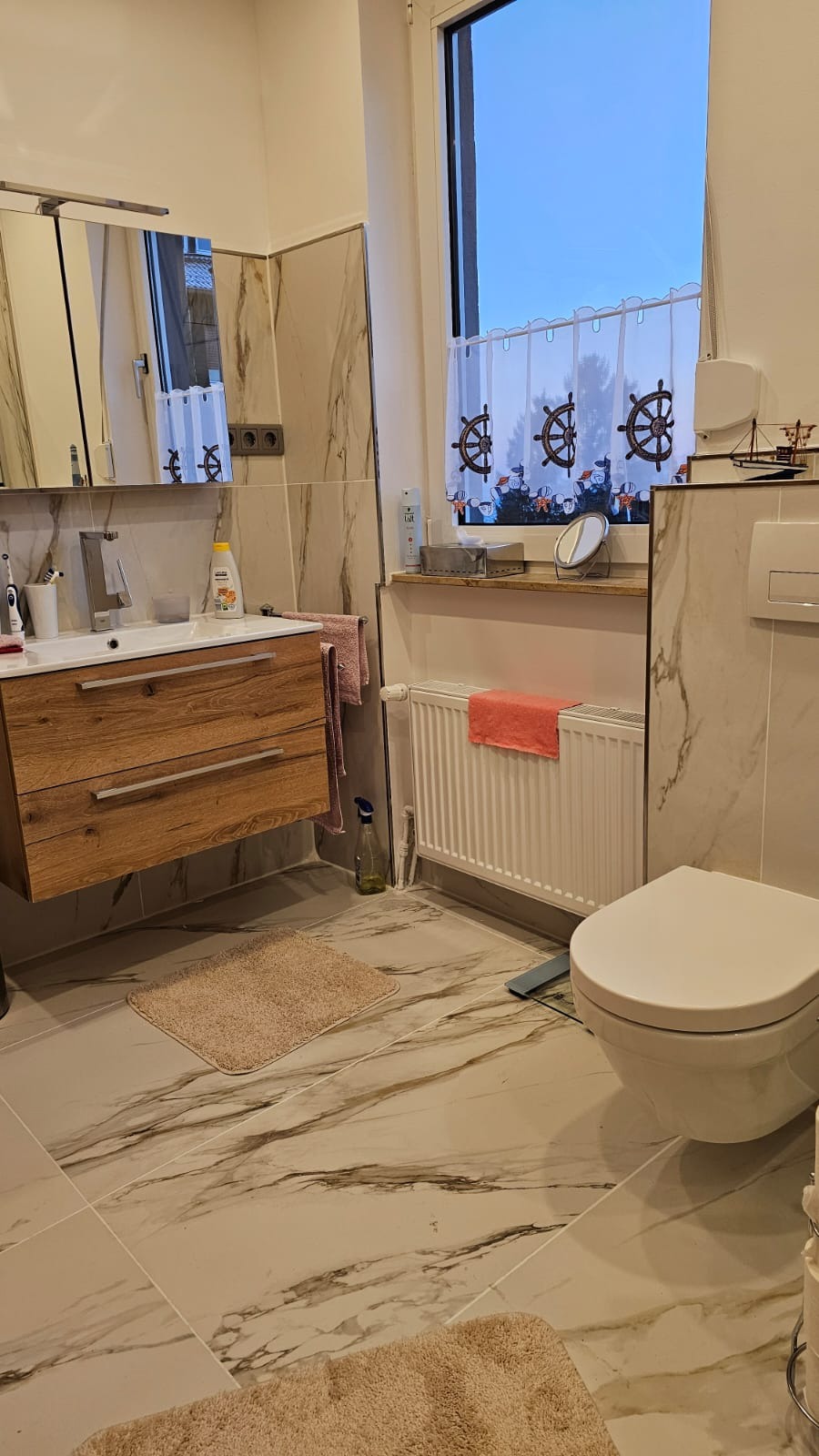 Wykończona łazienka w stylu marynistycznym: marmurowe płytki, drewniana szafka pod umywalką, okno z zasłonką w motywy żeglarskie, dekoracje z motywem statku.