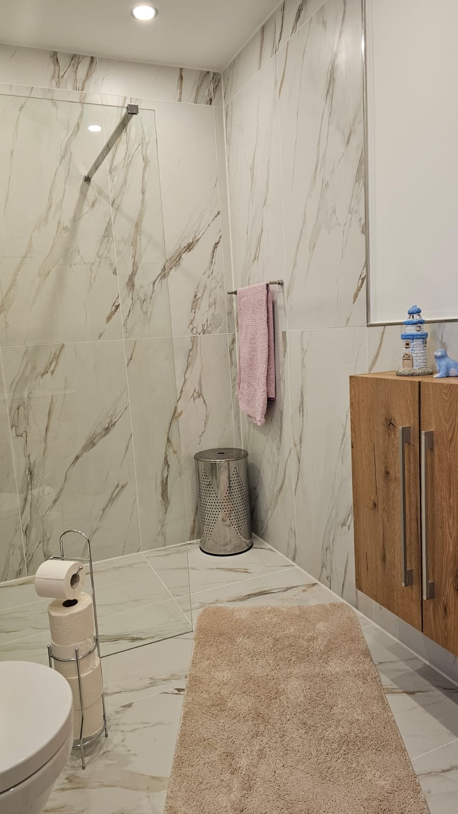 Elegancka łazienka z marmurowymi płytkami, kabiną prysznicową i drewnianą szafką. Różowy ręcznik i srebrny kosz na pranie dodają akcentu kolorystycznego.