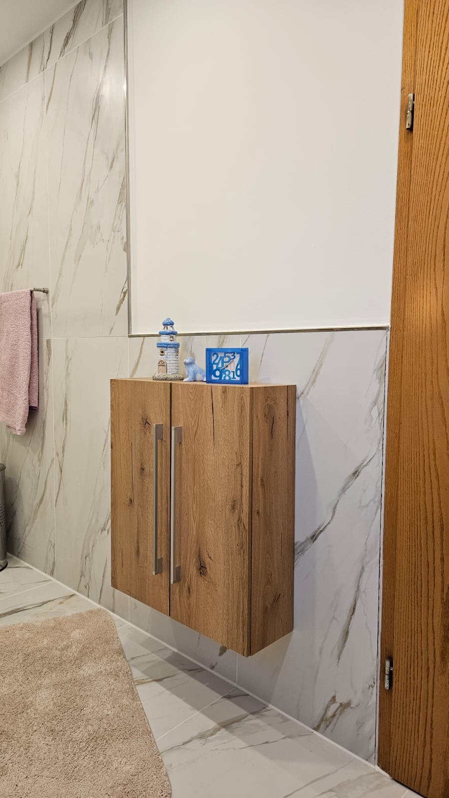 Nowoczesna łazienka z drewnianą szafką wiszącą, marmurowe płytki na ścianach i podłodze, dekoracje na szafce. Widok na fragment drzwi po prawej stronie.