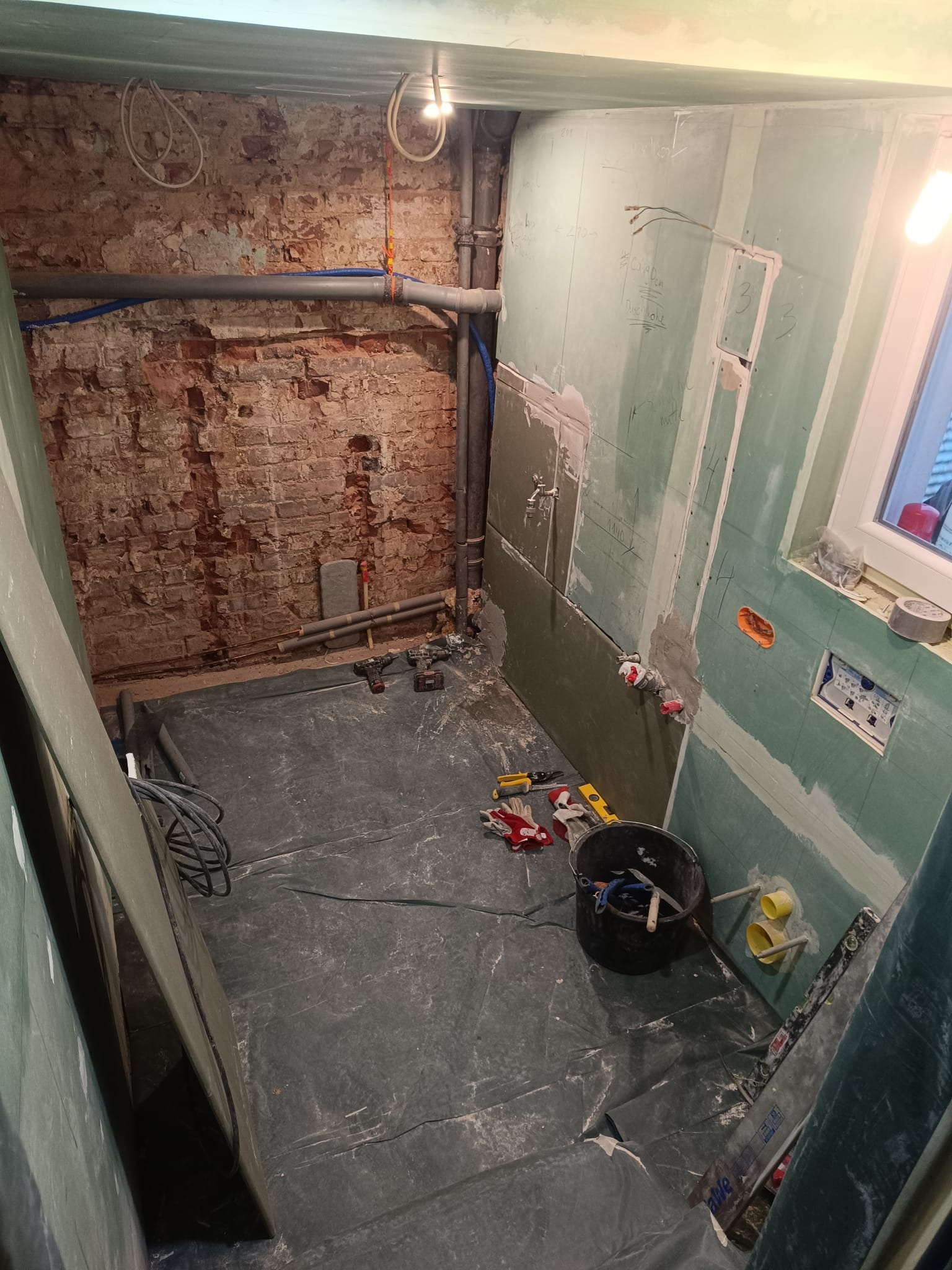 Wnętrze łazienki w trakcie remontu: surowa cegła, instalacje, płyty gipsowe, narzędzia i wiadro na podłodze. Widoczne przygotowania do wykończenia ścian.