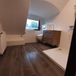 Ukończona łazienka na poddaszu w Jeleniej Górze z brązowymi płytkami imitującymi drewno na podłodze, biały brodzik, szafka z umywalką z drewnianymi szufladami oraz toaleta z widokiem na zieleń za...