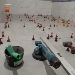 Wnętrze łazienki w trakcie układania płytek podłogowych, widoczne narzędzia glazurnicze: poziomica, wiadro z zaprawą, szlifierka kątowa oraz system poziomowania płytek.