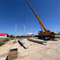 Montaż prefabrykowanych słupów betonowych za pomocą dźwigu na placu budowy, widoczni pracownicy w kamizelkach odblaskowych, w tle reklama Pucharu Narodów.