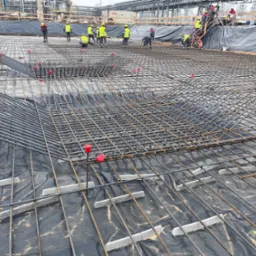 Zbrojenie fundamentu na czarnej folii z dystansami betonowymi, widoczna ekipa budowlana w żółtych kamizelkach i kaskach na tle instalacji przemysłowej.