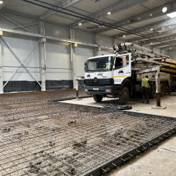 OPEN ACCESS SPÓŁKA Z OGRANICZONĄ ODPOWIEDZIALNOŚCIĄ - Wnętrze hali w trakcie budowy: zbrojenie pod wylewkę betonową, widoczny pracownik i betoniarka z pompą. Hala oświetlona, w tle ściany z płyt warstwowych.