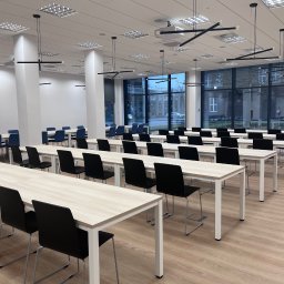 OPEN ACCESS SPÓŁKA Z OGRANICZONĄ ODPOWIEDZIALNOŚCIĄ - Jasne, przestronne wnętrze sali konferencyjnej z rzędami stołów i krzeseł, nowoczesnym oświetleniem i dużymi oknami. Widoczny projektor sufitowy.