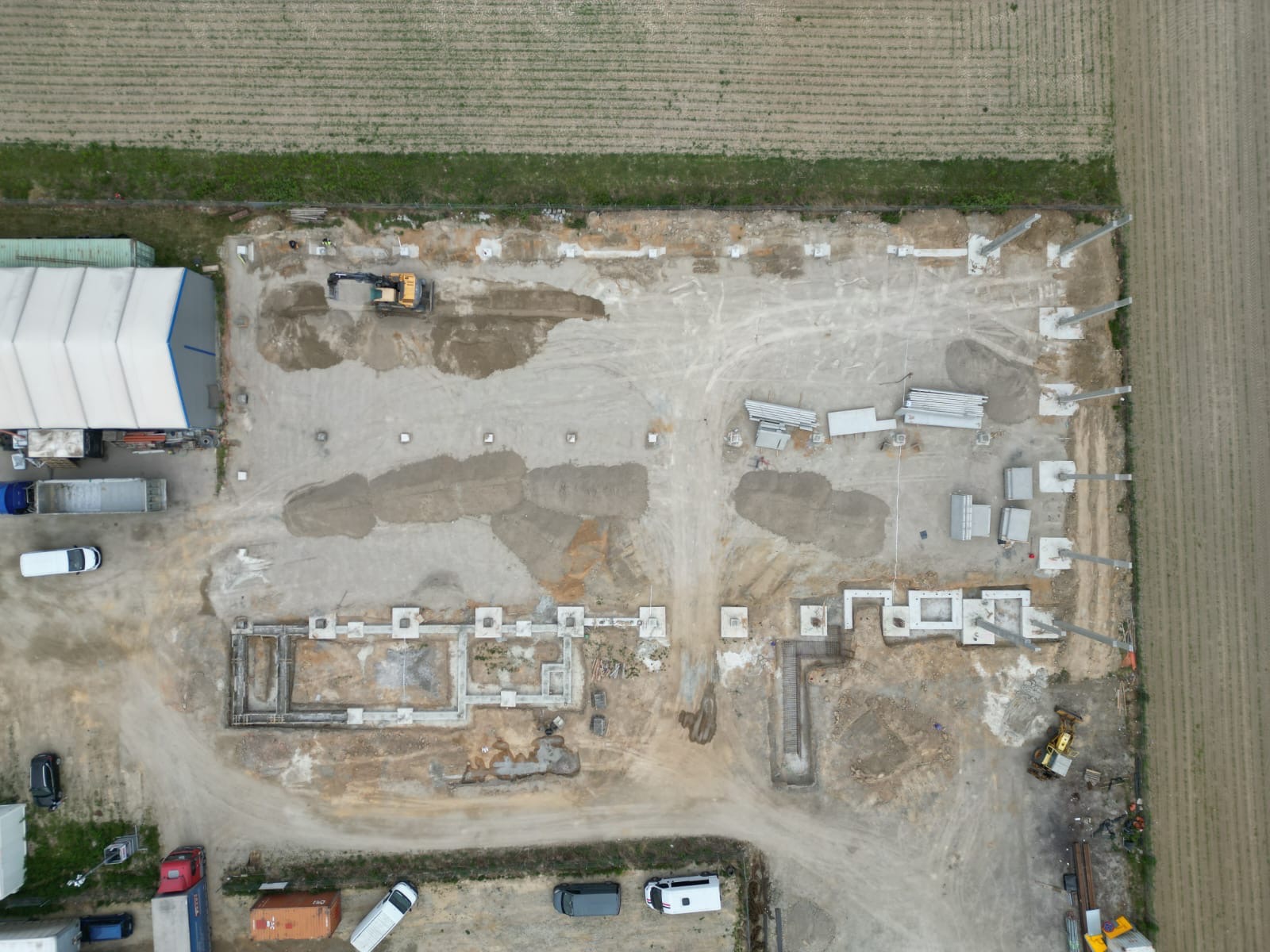 Widok z lotu ptaka na plac budowy z fundamentami, w tle pole uprawne i maszyny budowlane.
