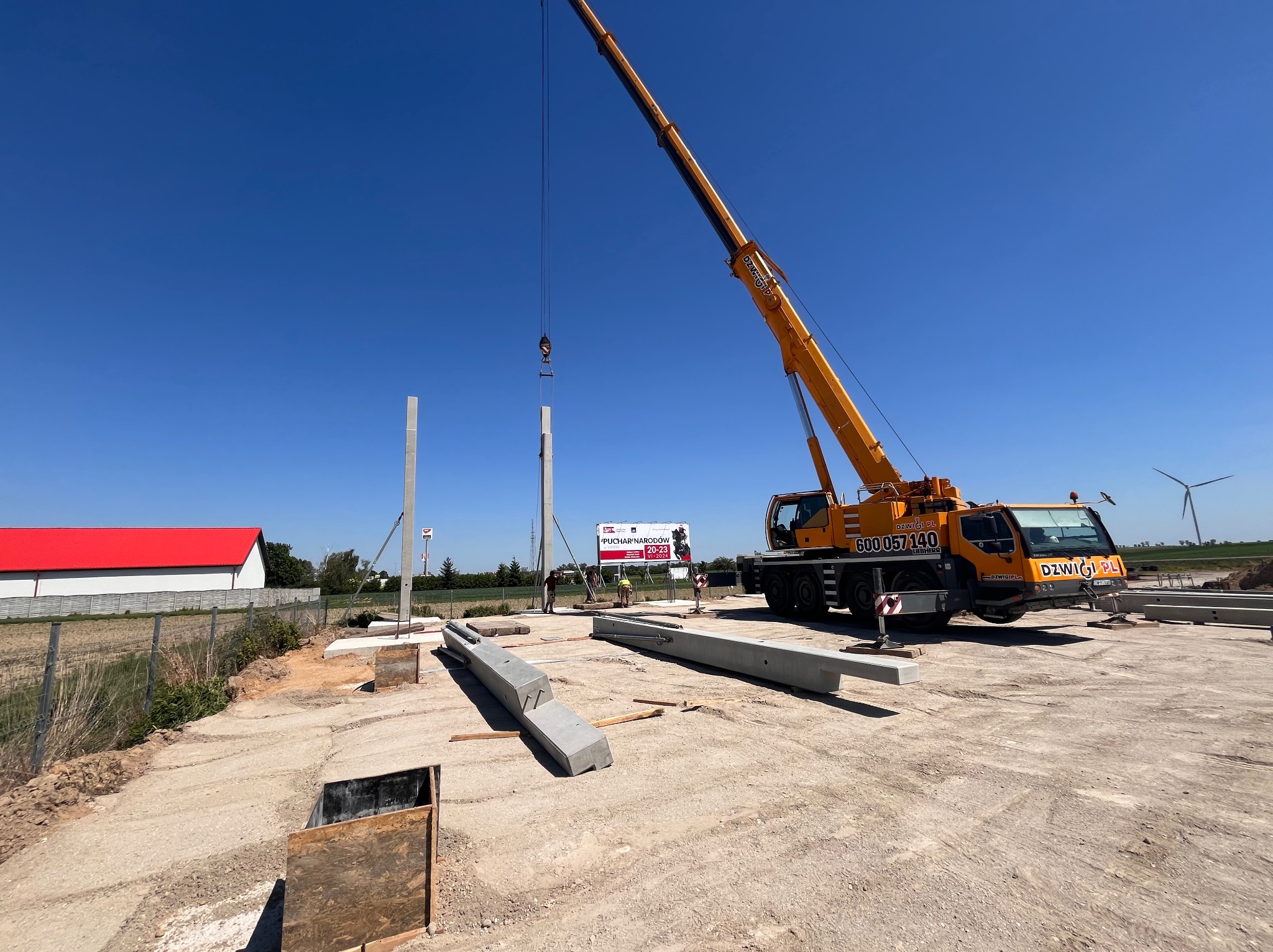 Montaż prefabrykowanych słupów betonowych za pomocą dźwigu na placu budowy, widoczni pracownicy w kamizelkach odblaskowych, w tle reklama Pucharu Narodów.