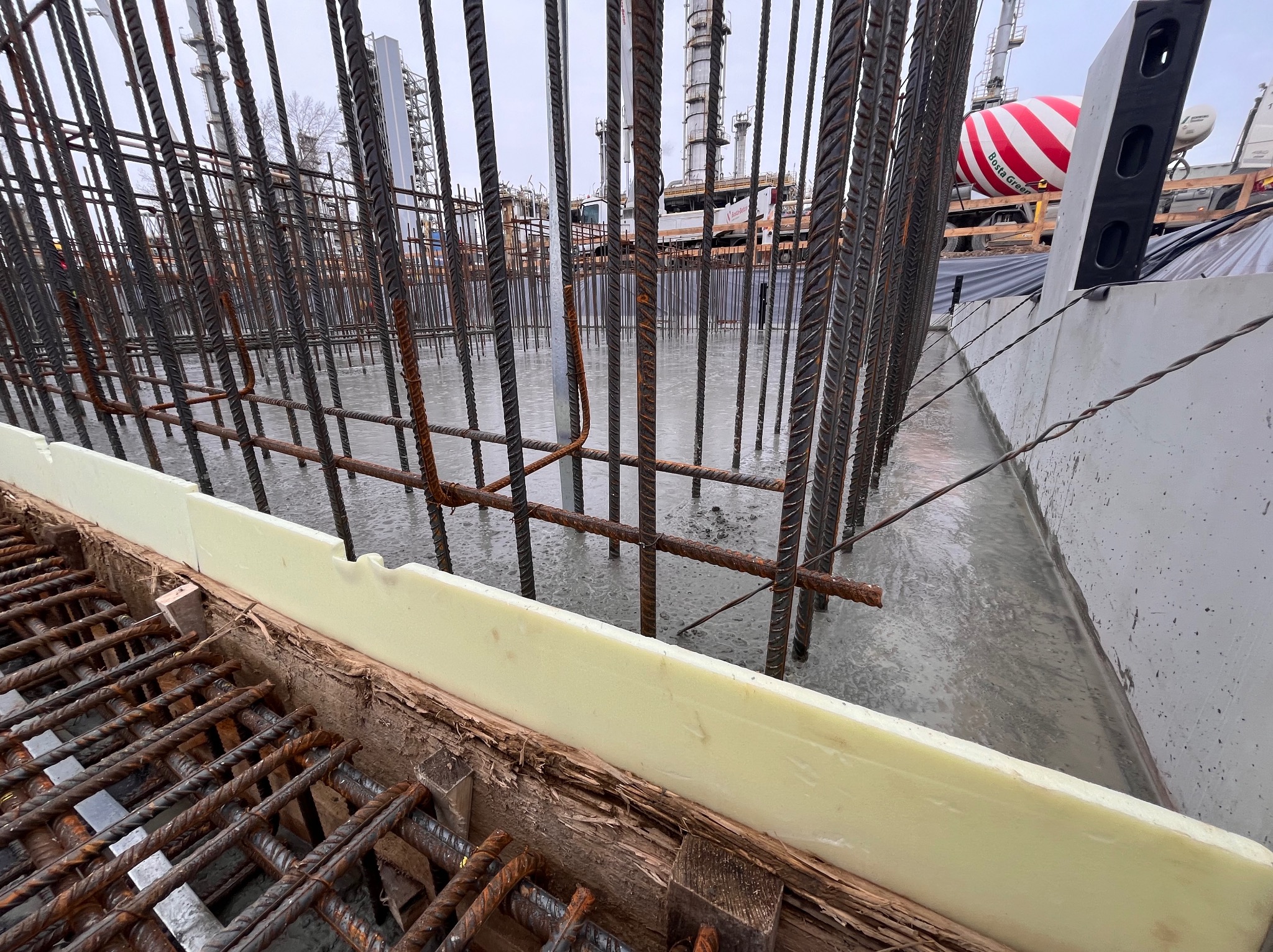 Zbrojenie fundamentu zalewane betonem, widoczna forma z izolacją, w tle betoniarka i elementy konstrukcji przemysłowej.