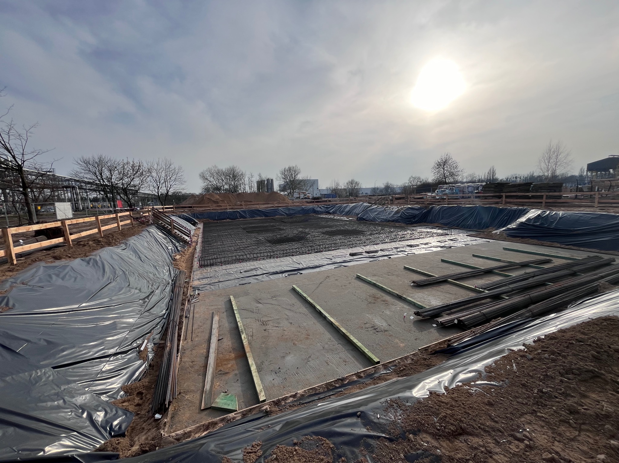 Przygotowanie fundamentów pod budynek przemysłowy: wykop wyłożony folią, zbrojenie siatką i drewniane szalunki, w tle zabudowania przemysłowe i pochmurne niebo.