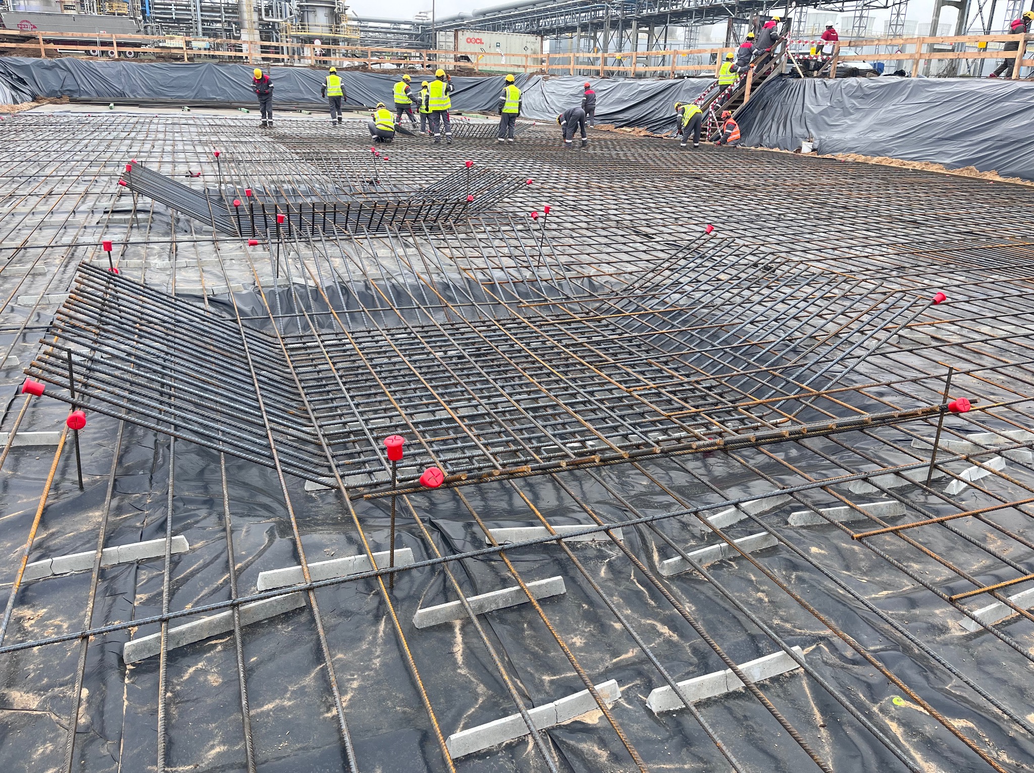 Zbrojenie fundamentu na czarnej folii z dystansami betonowymi, widoczna ekipa budowlana w żółtych kamizelkach i kaskach na tle instalacji przemysłowej.