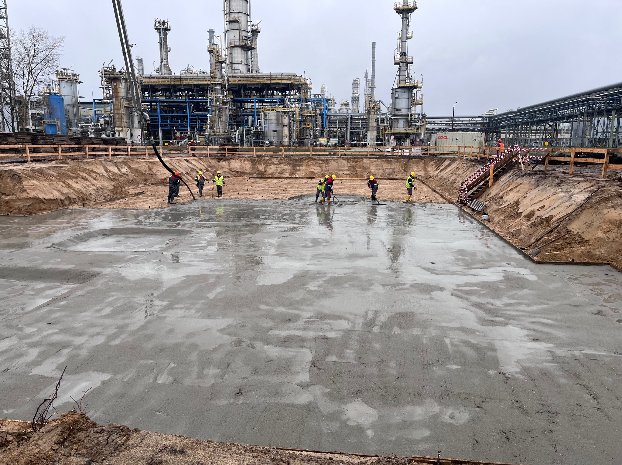 Robotnicy w kaskach i kamizelkach odblaskowych rozprowadzają świeżo wylany beton na placu budowy z kompleksem przemysłowym w tle.