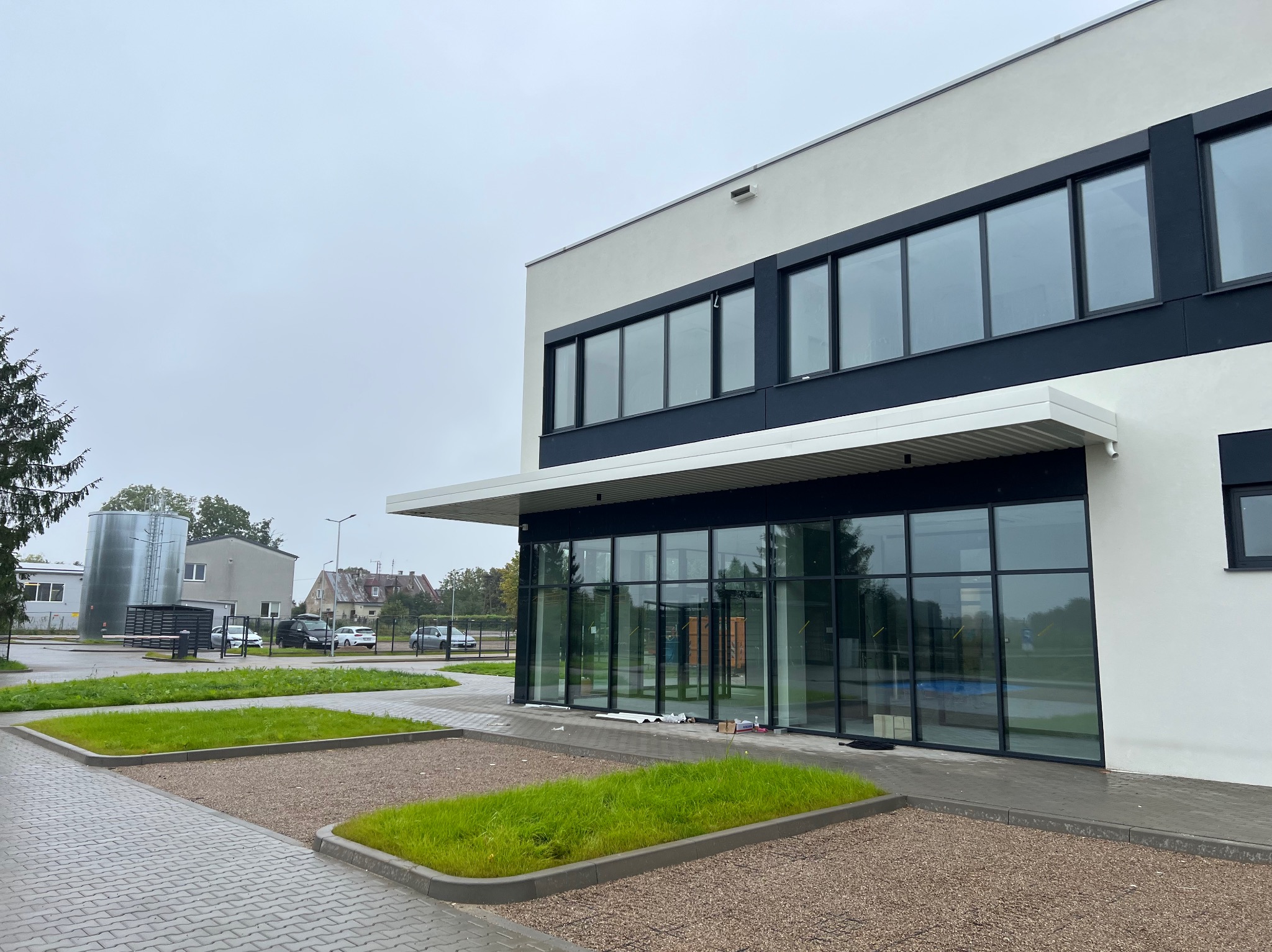 Nowoczesny budynek biurowy z dużymi oknami i minimalistyczną elewacją w kolorach bieli i czerni. Widoczne elementy zagospodarowania terenu i fragment sąsiedniej zabudowy.