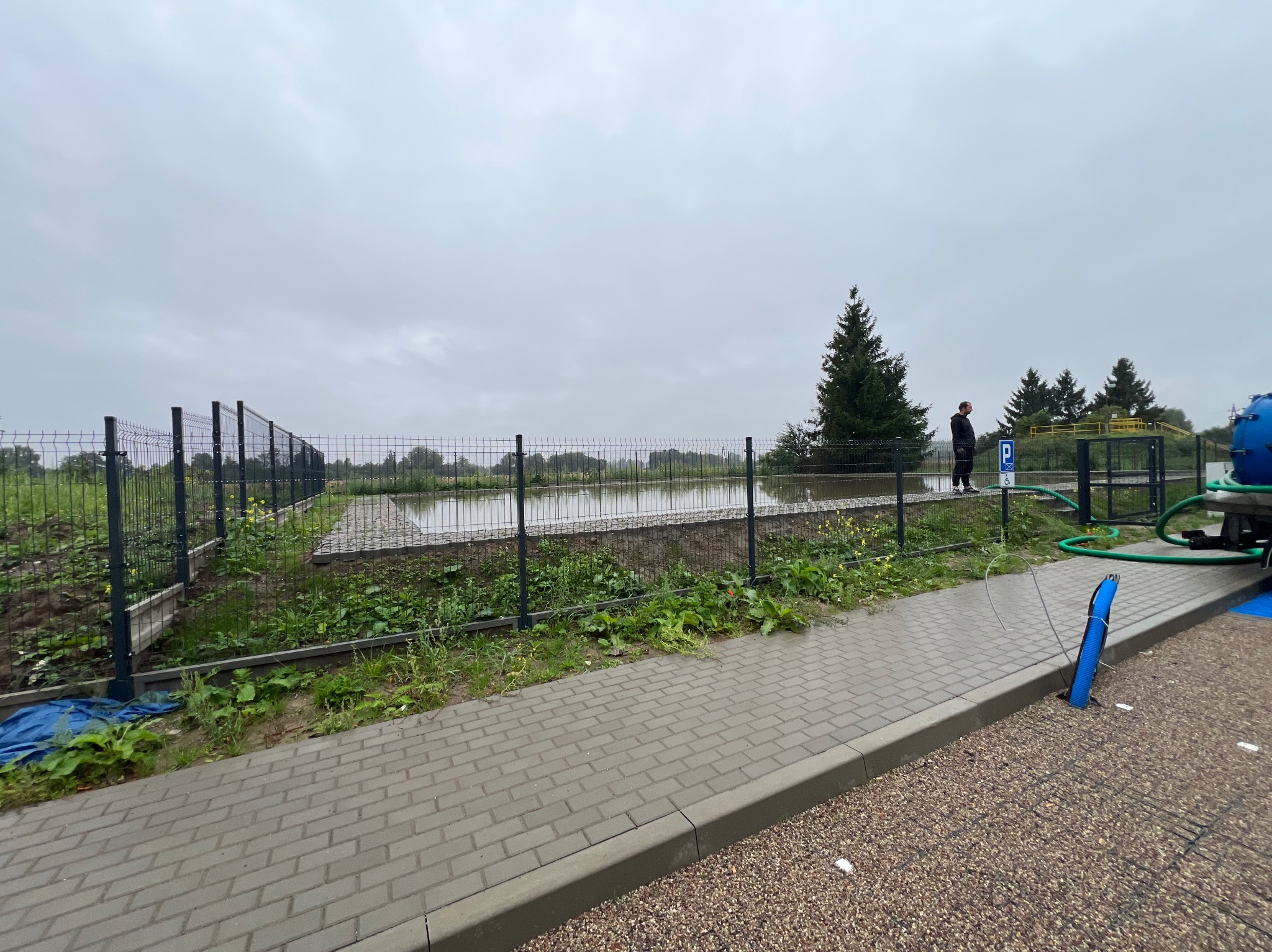 Zalany teren za ogrodzeniem z siatki, widoczny mężczyzna w oddali i fragment maszyny z wężami. Pochmurny dzień, mokra kostka brukowa.