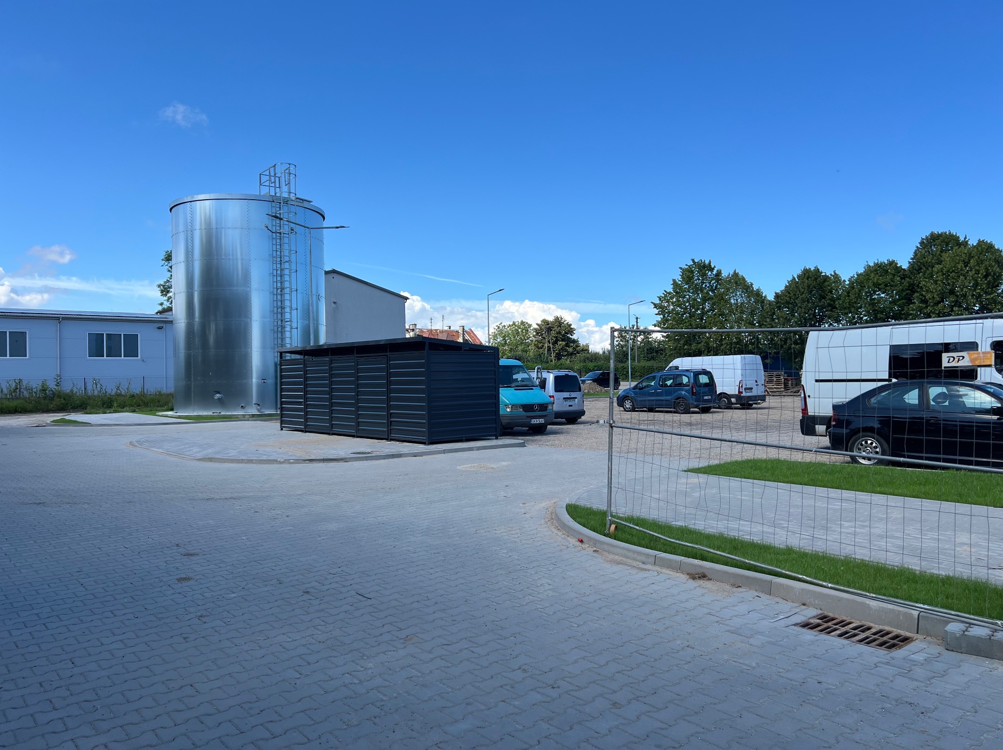 Srebrny zbiornik z drabinką obok budynku i czarnej wiaty na placu z kostki brukowej, w tle zaparkowane samochody i błękitne niebo.