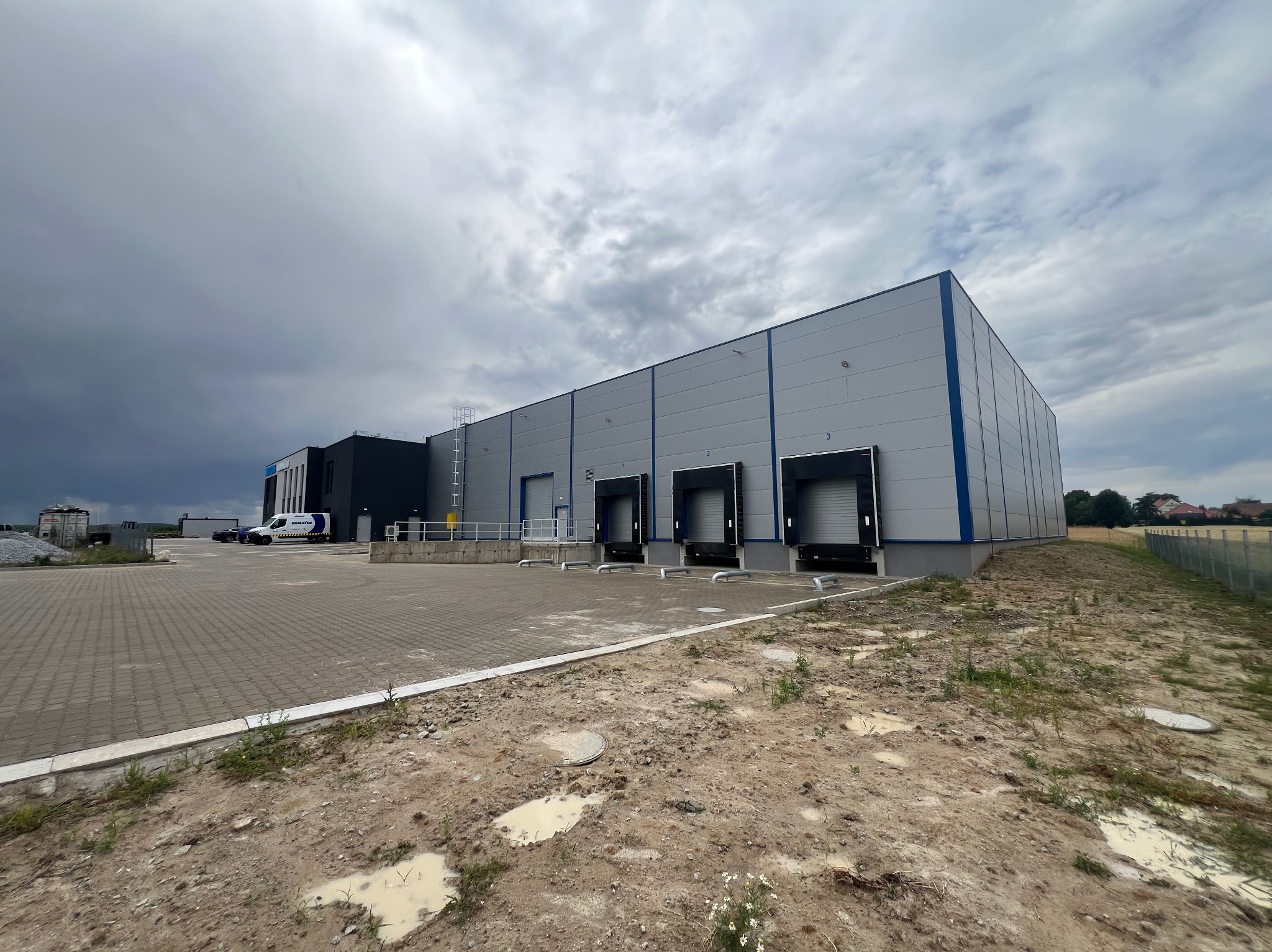 Nowoczesna hala magazynowa z rampami załadunkowymi, szara elewacja z niebieskimi akcentami, widok z zewnątrz na tle pochmurnego nieba i częściowo utwardzonego terenu.