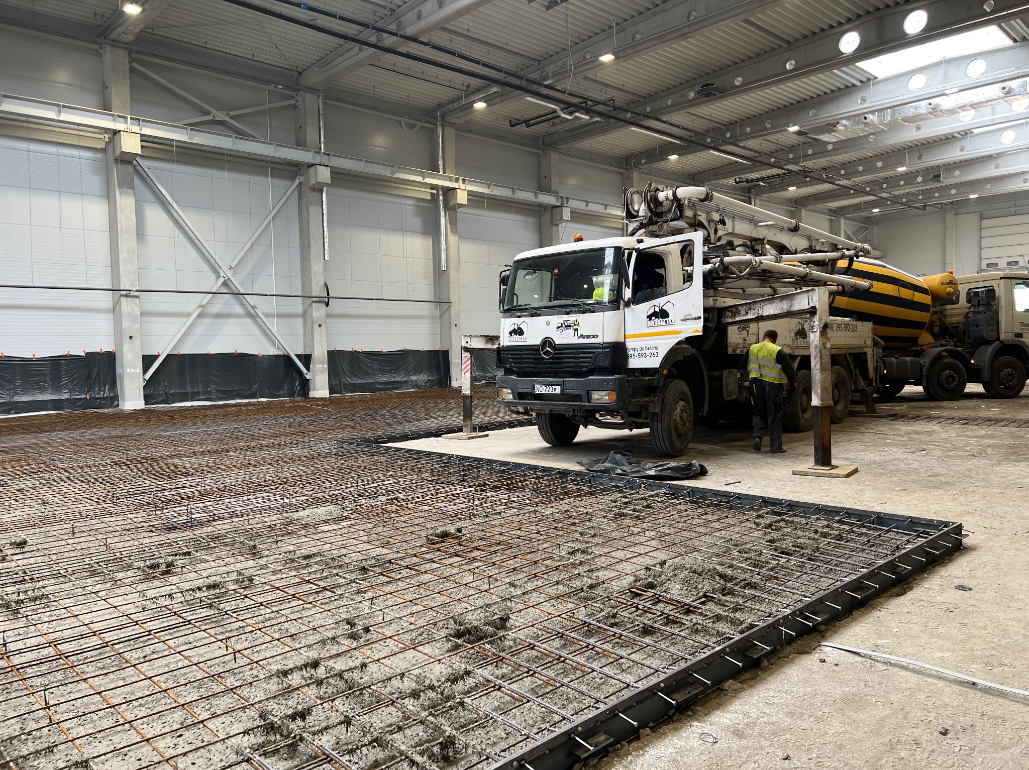 Wnętrze hali w trakcie budowy: zbrojenie pod wylewkę betonową, widoczny pracownik i betoniarka z pompą. Hala oświetlona, w tle ściany z płyt warstwowych.