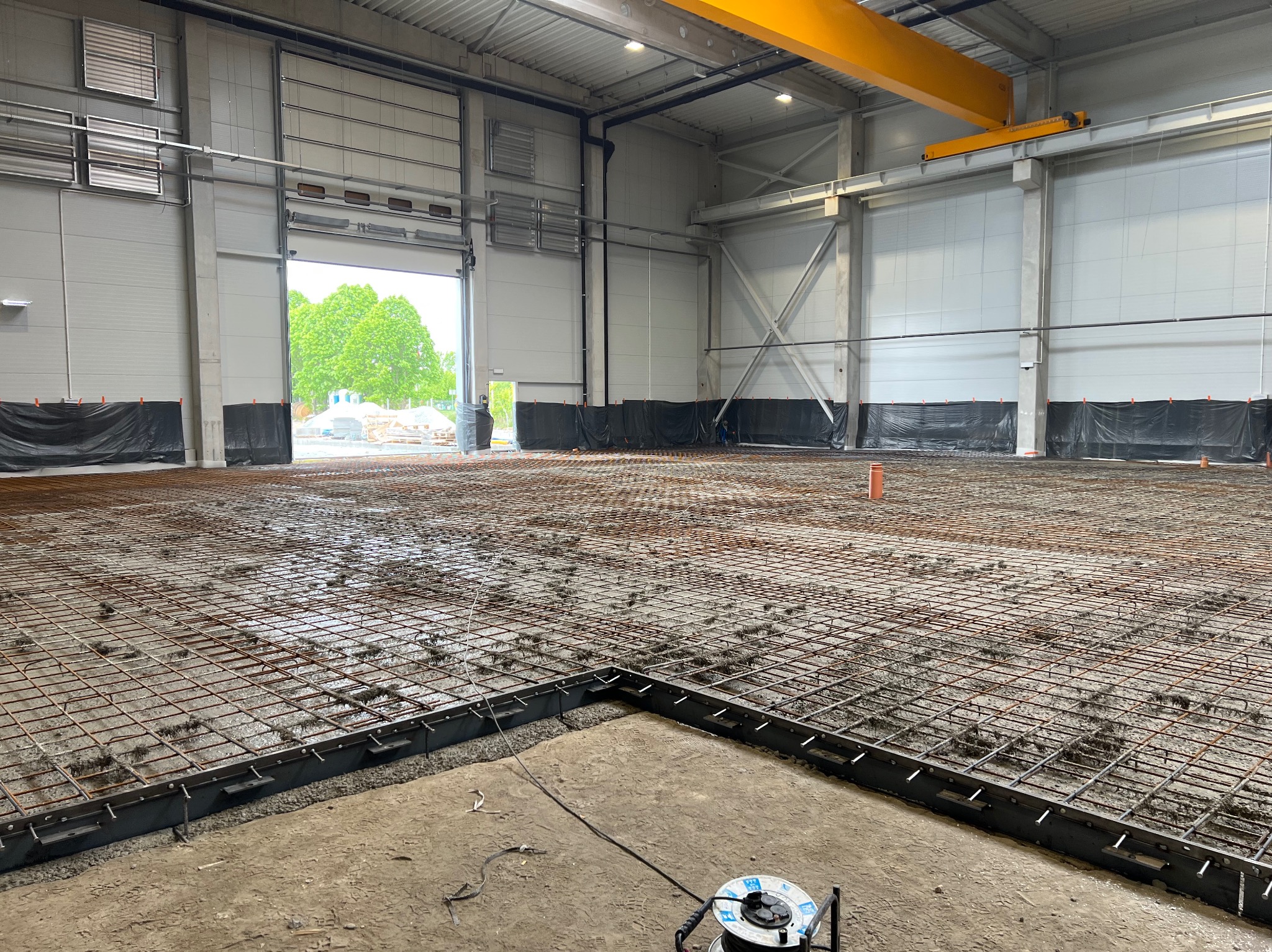 Zbrojenie pod wylewkę w hali przemysłowej, widoczne siatki zbrojeniowe, szalunki i częściowo otwarte wrota hali. Żuraw nad głową. Etap przygotowawczy do wylewania betonu.