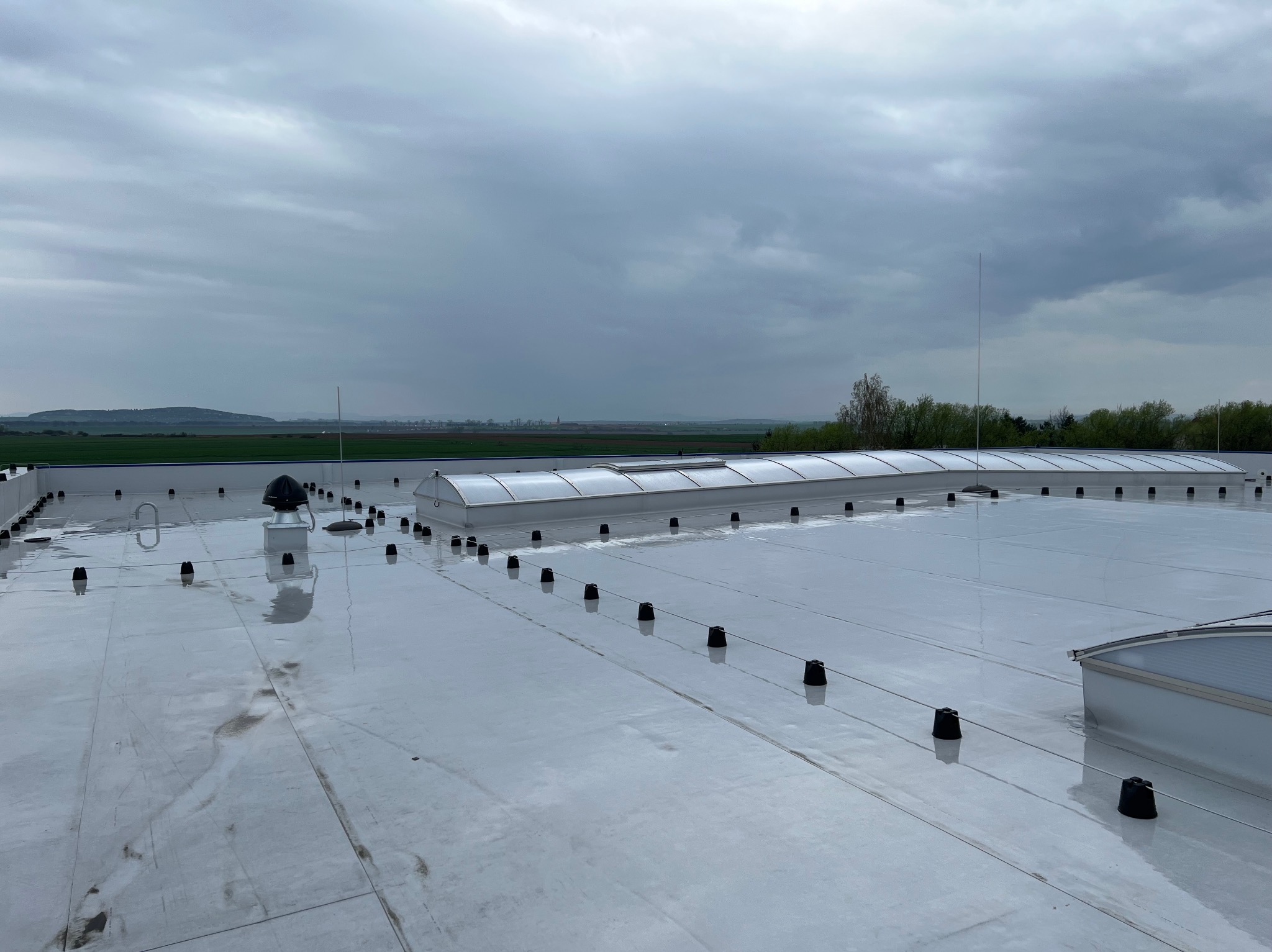 Biały dach hali przemysłowej z świetlikami i elementami mocującymi na tle pochmurnego nieba i zieleni. Widok z góry, lekko z boku.