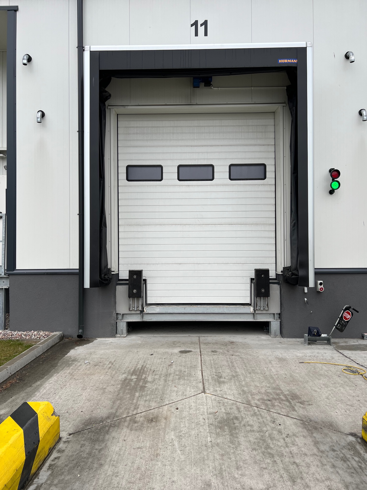 Biała brama segmentowa Hormann z trzema oknami w hali magazynowej, oświetlenie zewnętrzne, sygnalizacja świetlna, numer 11 nad bramą. Widok z zewnątrz na betonowy podjazd.