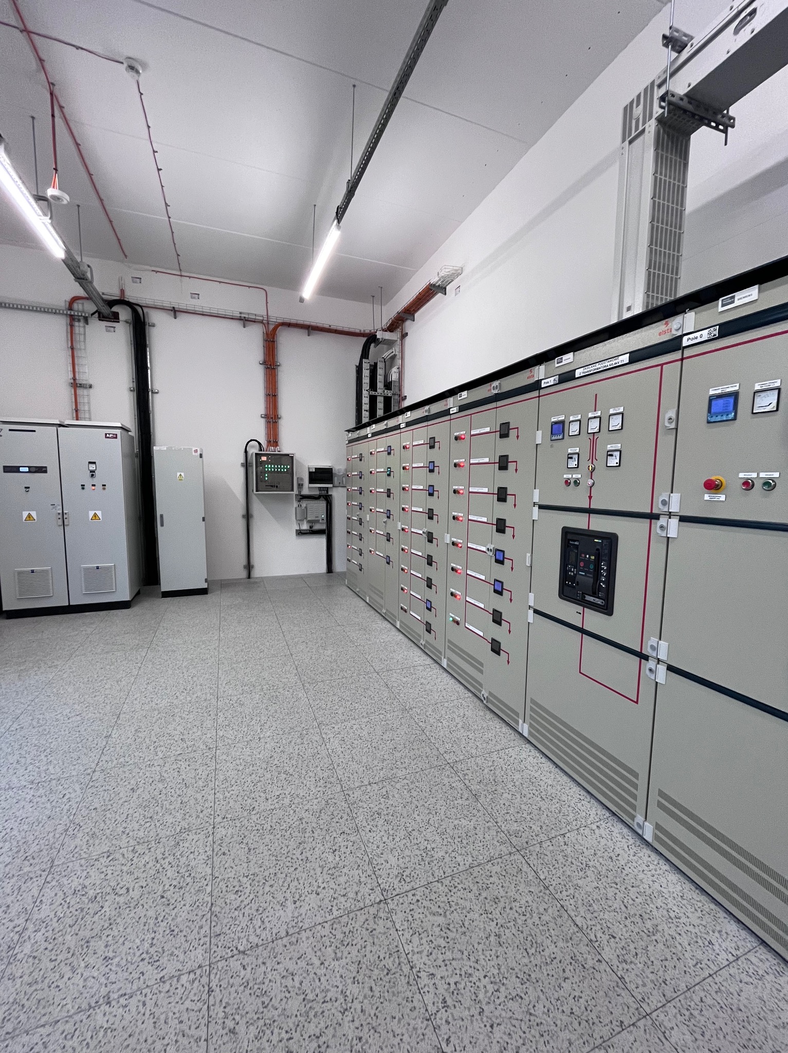 Szara rozdzielnica elektryczna w pomieszczeniu z szarą podłogą. Widoczne kable i rury na ścianach. Oświetlenie jarzeniowe.