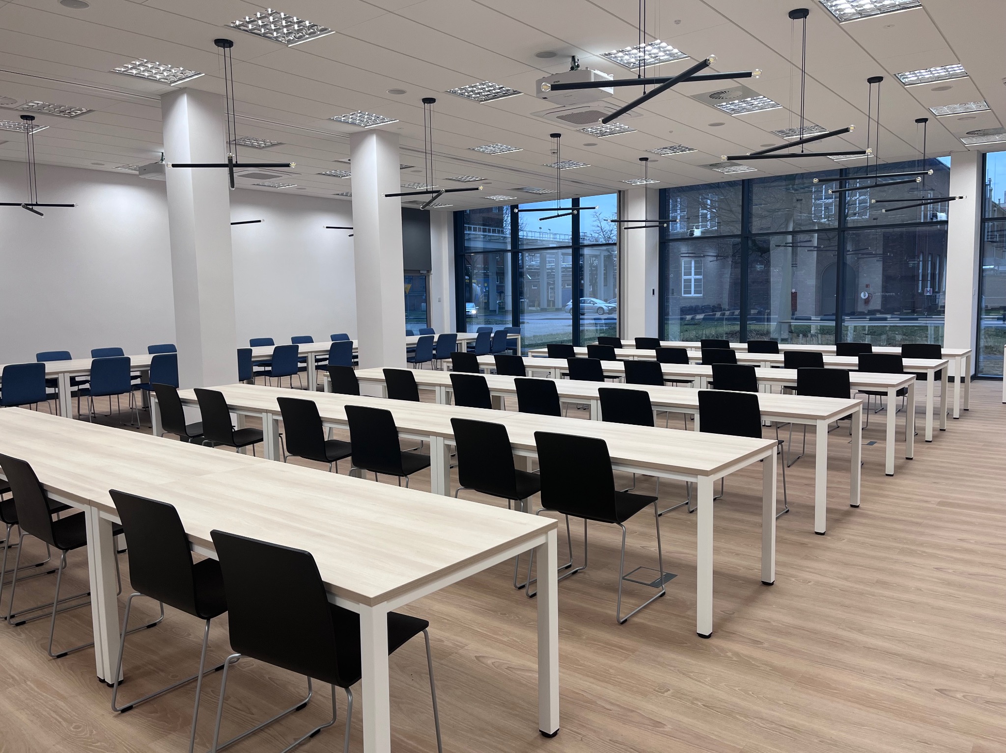 Jasne, przestronne wnętrze sali konferencyjnej z rzędami stołów i krzeseł, nowoczesnym oświetleniem i dużymi oknami. Widoczny projektor sufitowy.