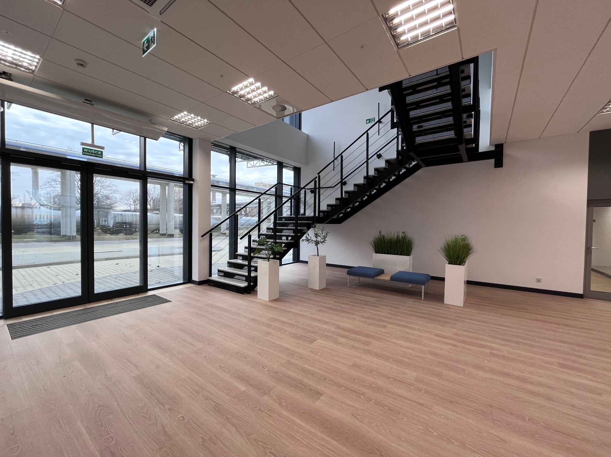 Nowoczesne wnętrze biurowe z drewnianą podłogą, schodami z czarną balustradą, dużymi oknami i roślinami w białych donicach. Widoczne elementy wykończenia i oświetlenie.