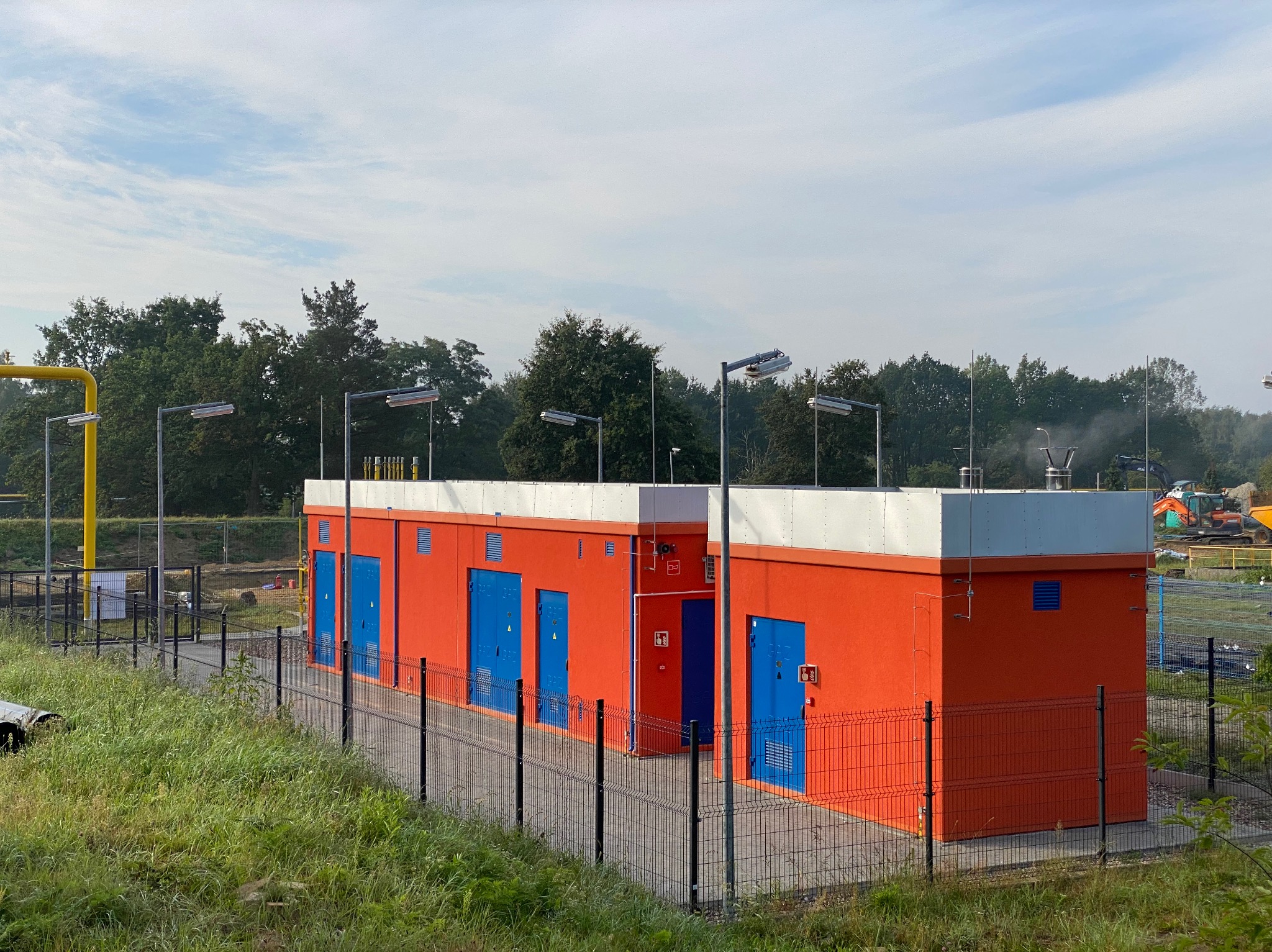 Pomarańczowy budynek stacji gazowej z niebieskimi drzwiami, otoczony metalowym ogrodzeniem. W tle widoczne drzewa i fragment żółtego rurociągu.