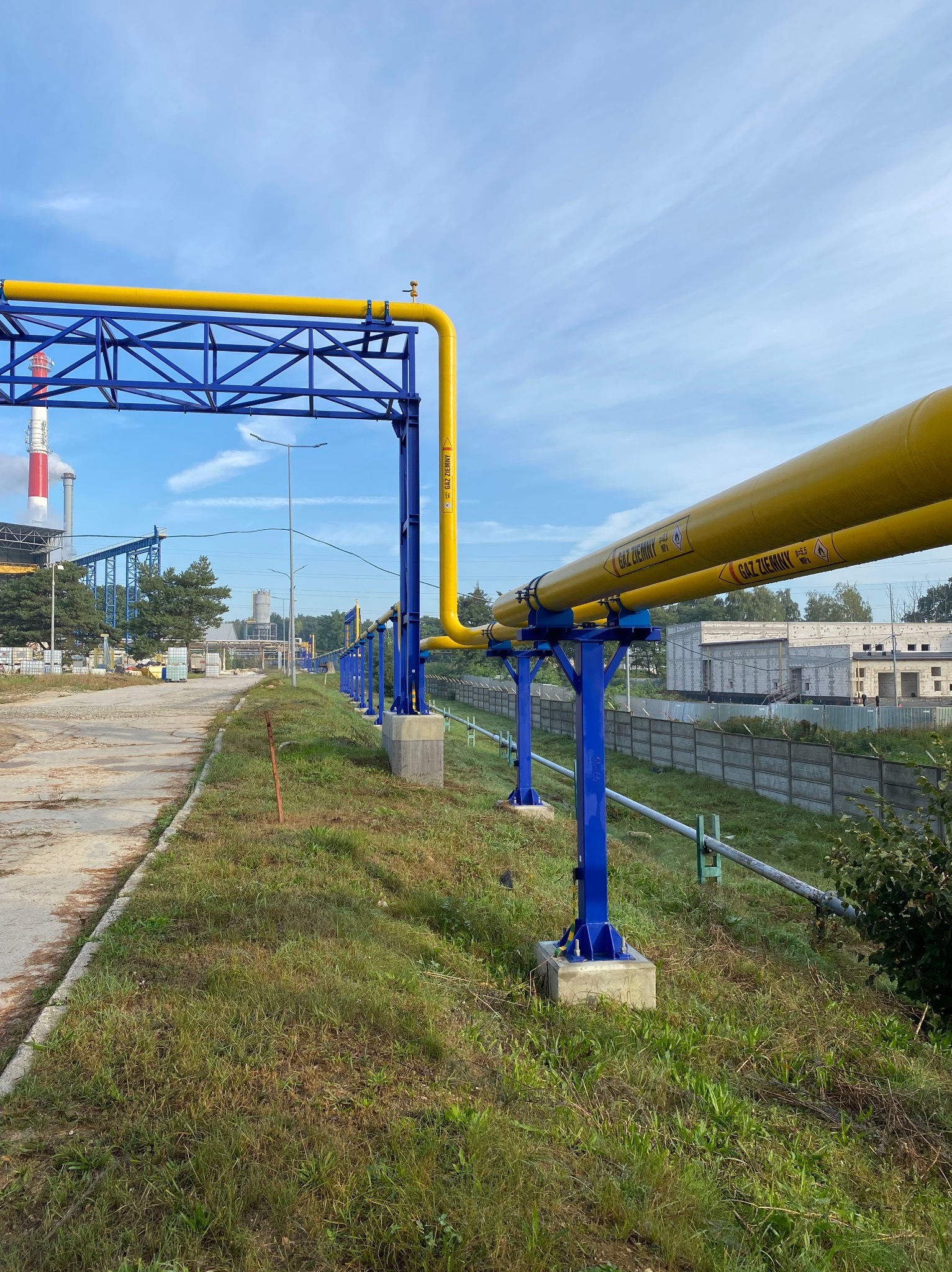 Żółte rury gazowe na niebieskiej konstrukcji wsporczej, z oznaczeniami ostrzegawczymi, na tle przemysłowego krajobrazu i błękitnego nieba.