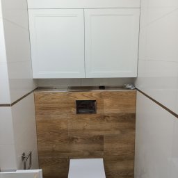 MAG-DREW - Wnętrze łazienki w trakcie remontu: białe płytki, drewniana zabudowa ściany z ukrytą spłuczką, wisząca toaleta z folią ochronną, białe szafki nad zabudową, widoczne elementy montażowe szafki...