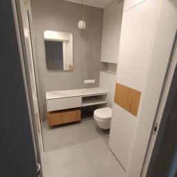 MAG-DREW - Nowoczesna łazienka z szarą ścianą, szafką pod umywalką z drewnianymi akcentami, białymi szafkami wiszącymi i podwieszanym WC, oświetlona złotą lampą wiszącą.