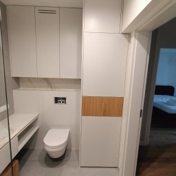 MAG-DREW - Nowoczesna łazienka z zabudową meblową na wymiar w odcieniach bieli i drewna, podwieszana toaleta i umywalka z blatem, widok na sypialnię przez otwarte drzwi.