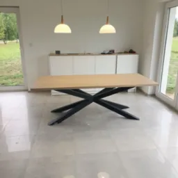 Stół z blatem w odcieniach drewna i geometryczną, czarną podstawą w minimalistycznym, jasnym wnętrzu z dwiema lampami wiszącymi i szafką w tle.