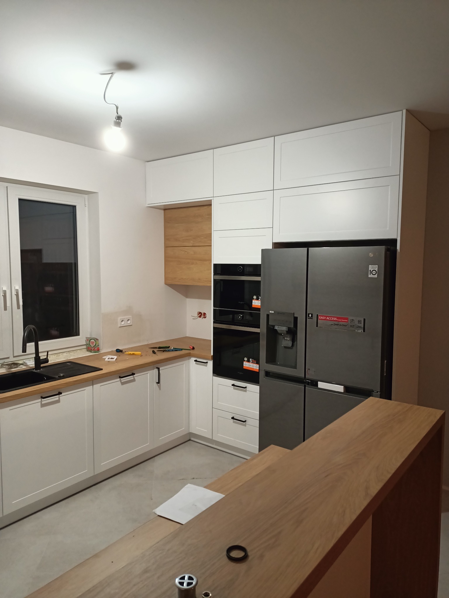 Nowoczesna kuchnia w trakcie montażu: białe szafki z drewnianymi akcentami, czarny zlew, stalowa lodówka side-by-side, widoczne narzędzia na blacie.