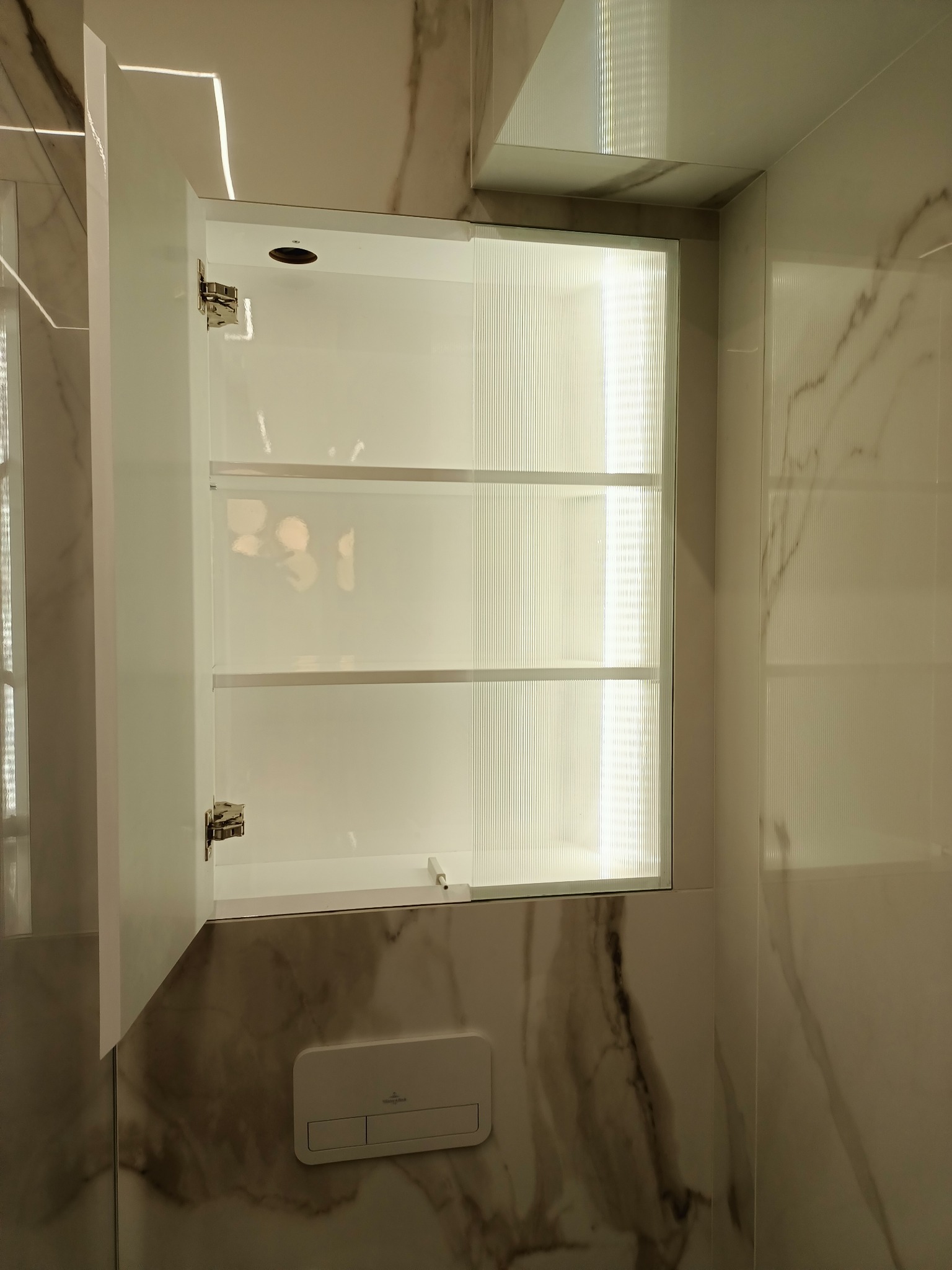 Biała szafka wnękowa z oświetleniem LED wewnątrz, zamontowana w łazience wykończonej marmurkowymi płytkami. Drzwiczki szafki otwarte, widoczne trzy półki. Na ścianie widoczny panel spłuczki WC.