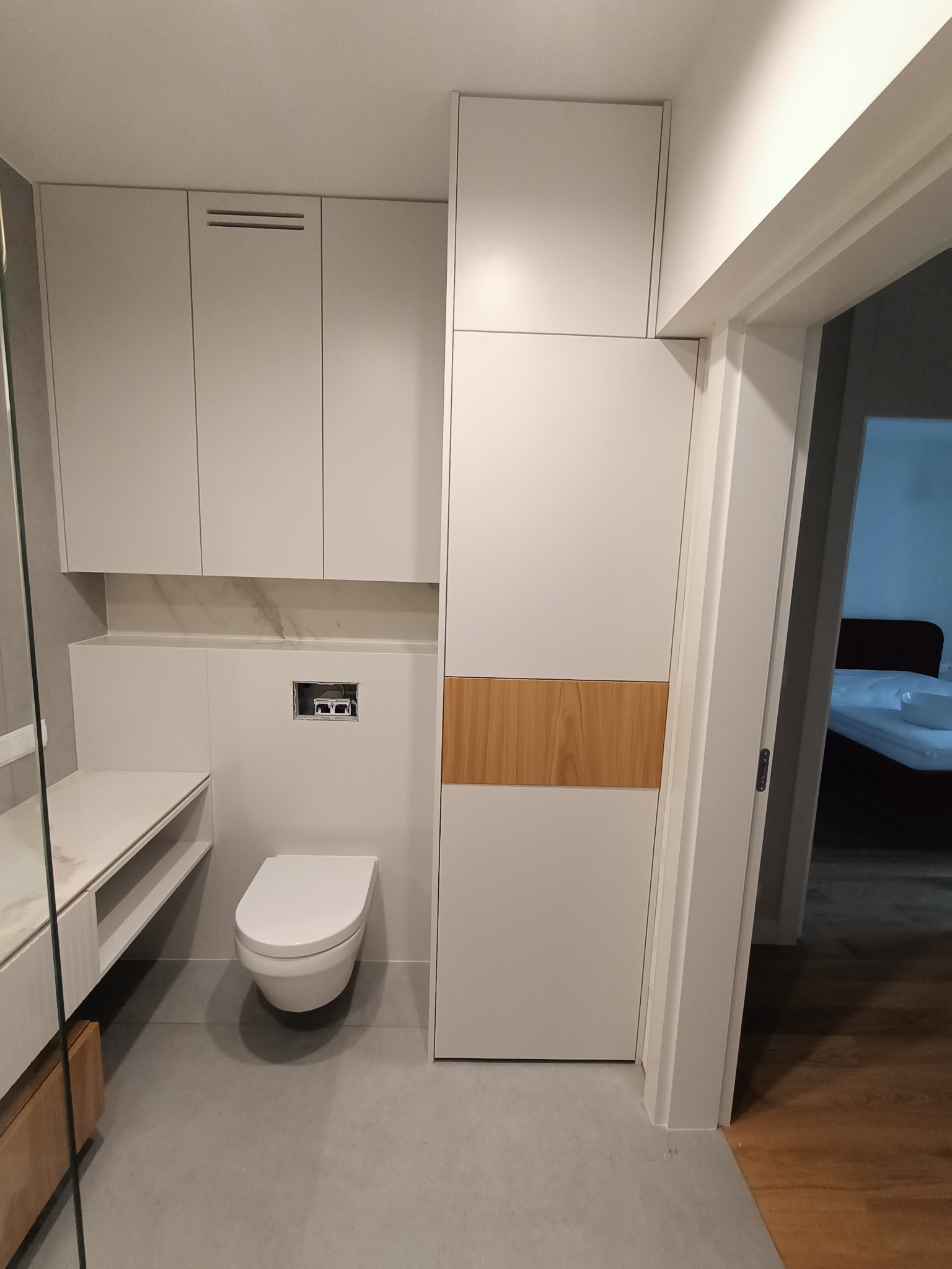 Nowoczesna łazienka z zabudową meblową na wymiar w odcieniach bieli i drewna, podwieszana toaleta i umywalka z blatem, widok na sypialnię przez otwarte drzwi.