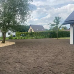 Przygotowany teren pod trawnik, świeżo wyrównana ziemia z widocznymi śladami opon, fragment wysypanego piasku, w tle domy jednorodzinne i zielony żywopłot.