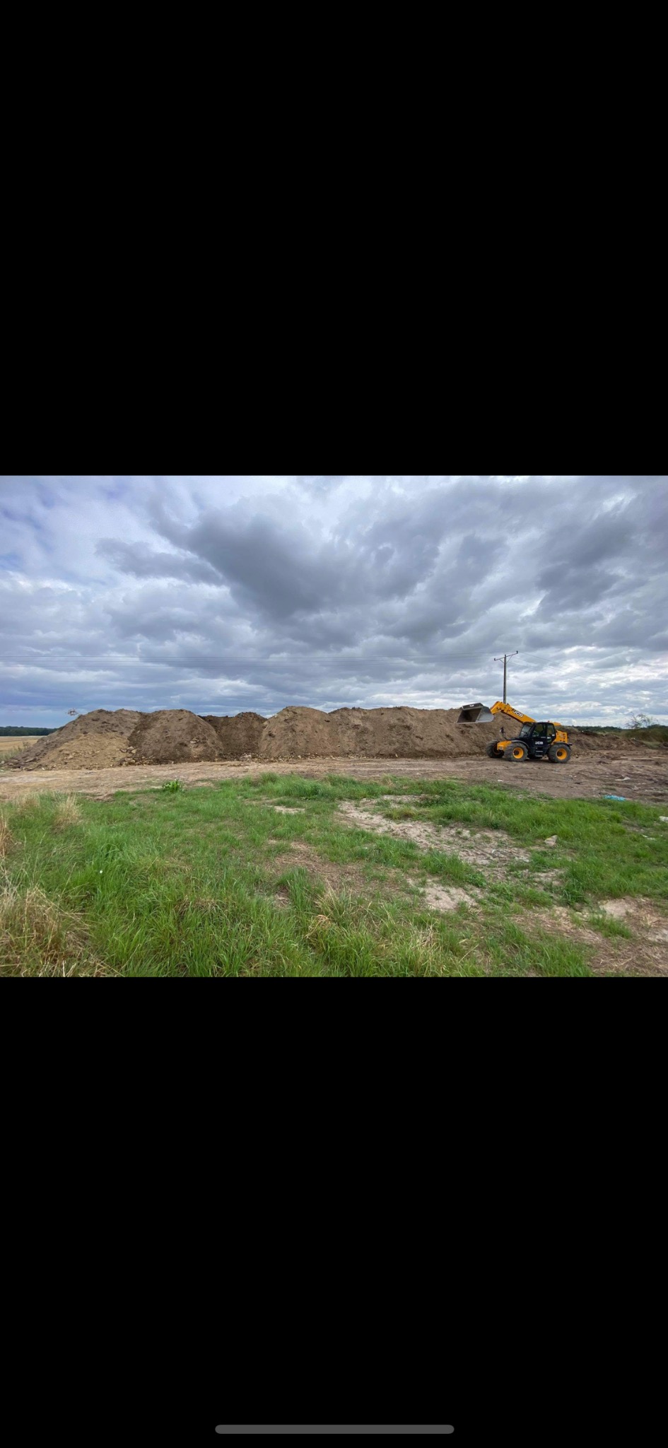Żółta ładowarka teleskopowa stoi obok pryzmy ziemi na placu budowy, pochmurne niebo w tle.