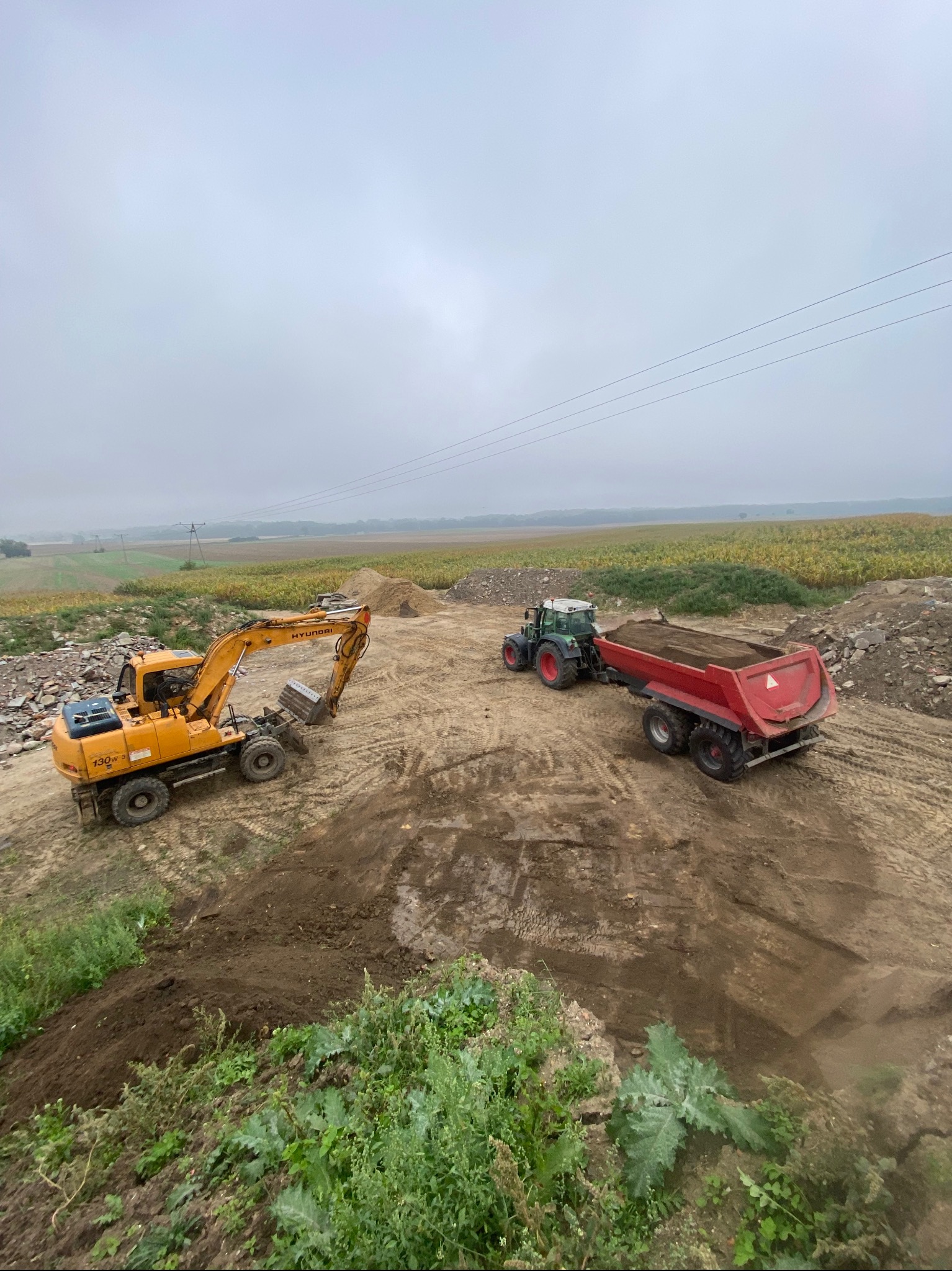 Żółta koparka Hyundai 130W-3 i ciągnik z czerwoną przyczepą na placu budowy, w tle pole i pochmurne niebo.