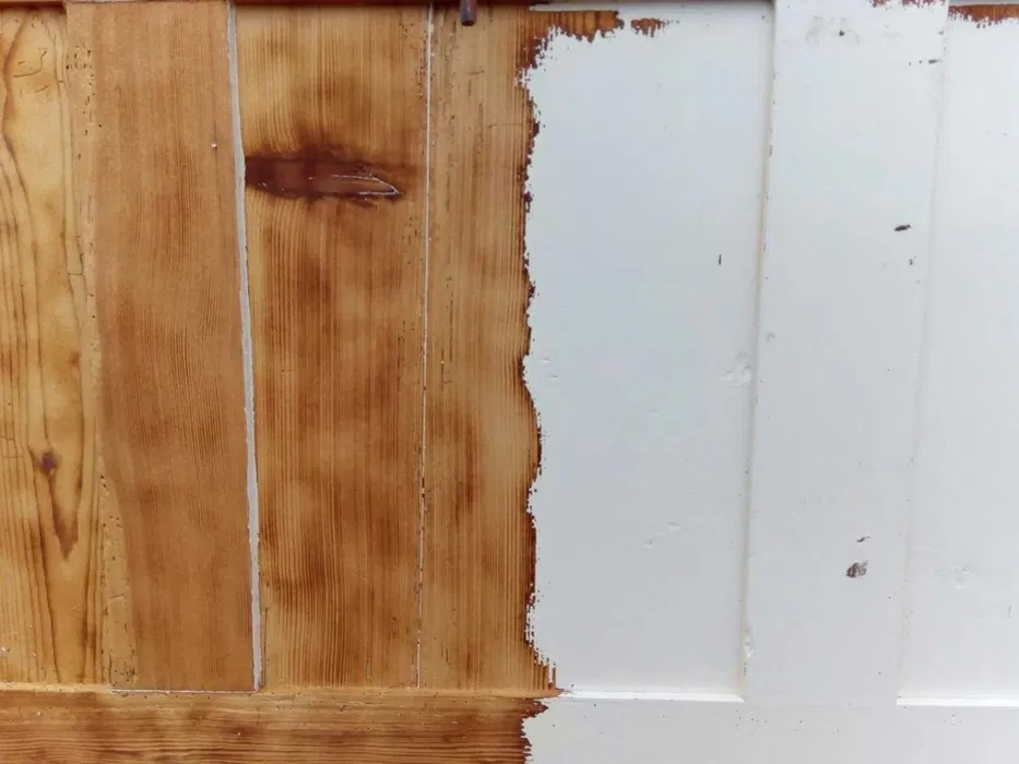 Częściowo pomalowane drewniane drzwi z widoczną strukturą drewna po lewej stronie i świeżą, białą farbą po prawej stronie, ukazujące proces renowacji.