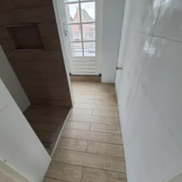 Wykończona łazienka z drewnopodobnymi płytkami na podłodze i ścianach, białe płytki na ścianie, widoczny odpływ liniowy i okno z widokiem na budynki.