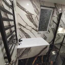 Robert Myslinski - Nowoczesna łazienka z białą wanną obudowaną płytkami imitującymi marmur, czarnym grzejnikiem drabinkowym i podłogą z czarnego marmuru z złotymi żyłami. Uwagę przyciąga okno ze wzorem kwiatowym.